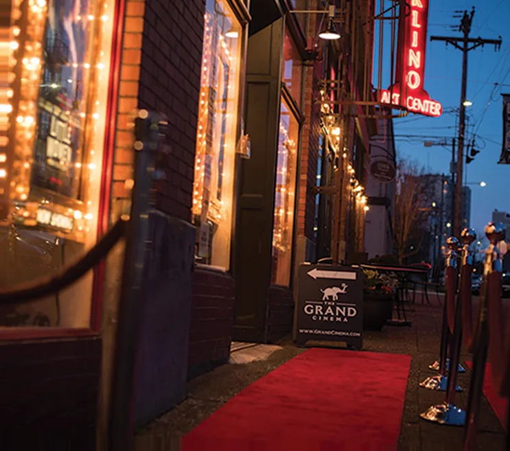 Evening sidewalk outside The Grand Cinema, with a red carpet, stanchions, string lights in the windows and a sandwich board for the theater.