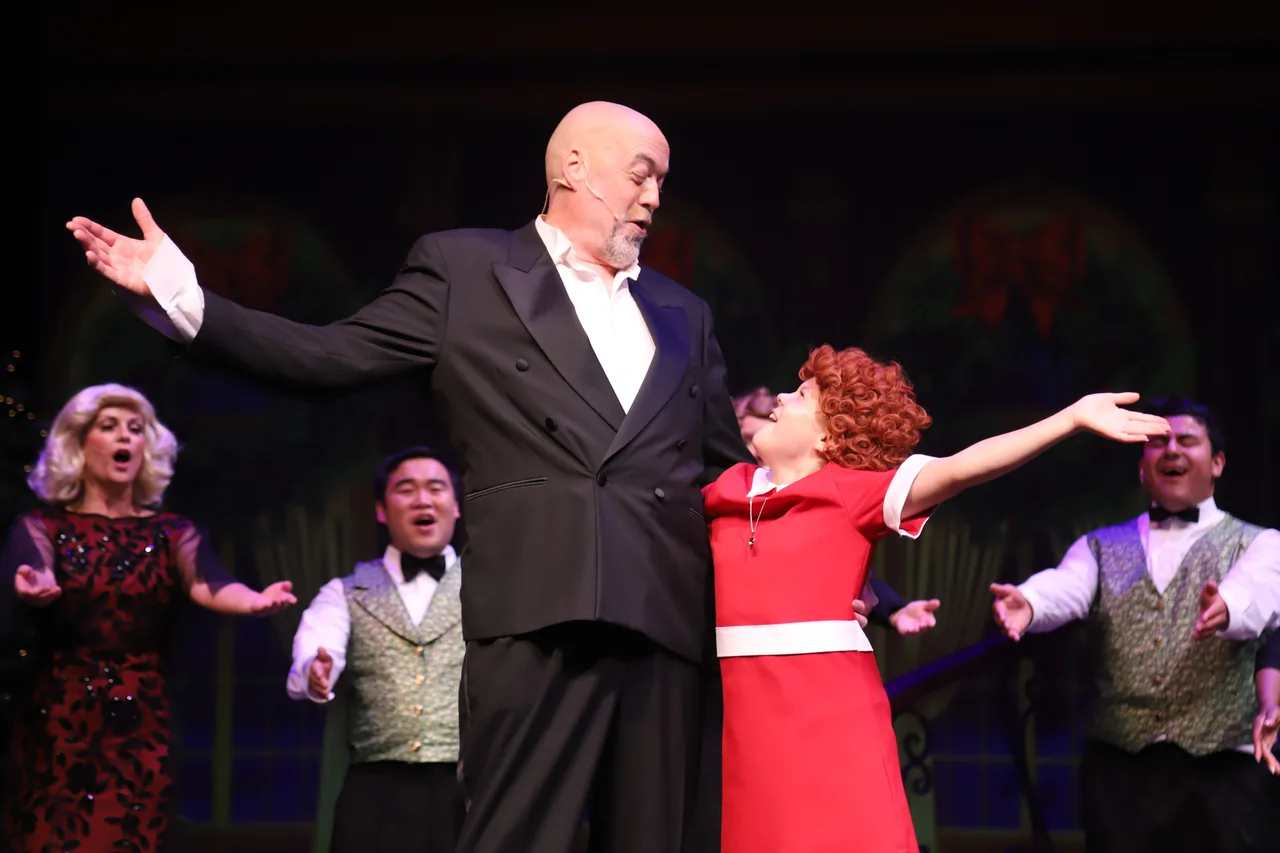 A bald man in a tuxedo and a woman in a red dress stand center stage with arms open, ensemble singers behind them.